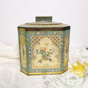 May include: A hexagonal metal storage tin featuring a light blue and cream color palette with intricate floral patterns, a central butterfly motif, and a geometric lattice border. The container includes a matching lid with a rectangular handle.
