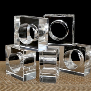 Puede incluir: Conjunto de cinco servilleteros de acrílico transparente. Cada servilletero tiene forma de cubo con una abertura circular en el centro. Están dispuestos sobre una superficie de madera sobre un fondo negro.