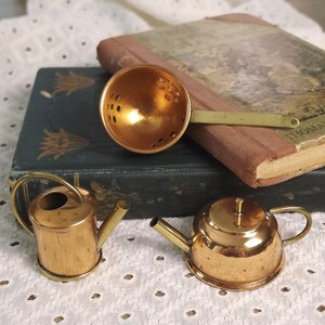 Könnte beinhalten: Ein Teesieb aus Messing mit perforierter Schale, eine kleine Gießkanne aus Messing und eine Miniatur-Teekanne aus Messing. Die Gegenstände liegen neben einem dunkelgrünen Buch mit goldenen Blumenmustern und einem hellbraunen Buch mit Illustration.