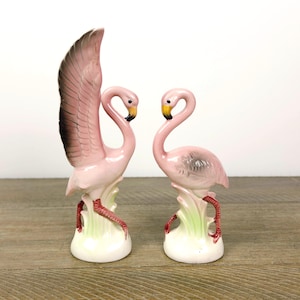 Puede incluir: Dos figuras de flamencos de cerámica. Un flamenco tiene un ala levantada con detalles negros y marrones. Ambos flamencos son rosas con picos amarillos y patas rojas, de pie sobre una base blanca con detalles verdes. Decorativo.