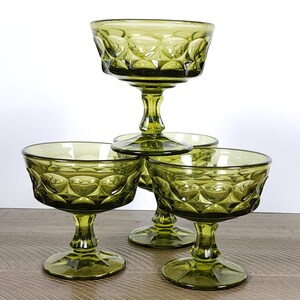 Puede incluir: Cuatro copas de champán vintage de color verde oliva. Las copas tienen un patrón de diamantes en el cuenco y una base de pedestal. Las copas están apiladas en forma de pirámide sobre una superficie de madera.