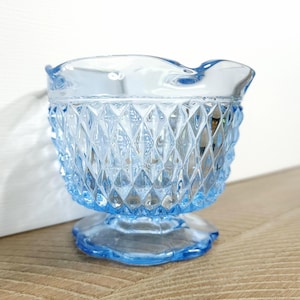 May include: A small, light blue glass bowl with a diamond pattern and a scalloped rim. The bowl has a short, flared foot.