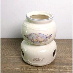 May include: A two-piece ceramic warmer featuring an off-white speckled finish. The top section displays a central oval illustration of two geese adorned with pink ribbons, surrounded by pink roses. The base has two circular side openings.