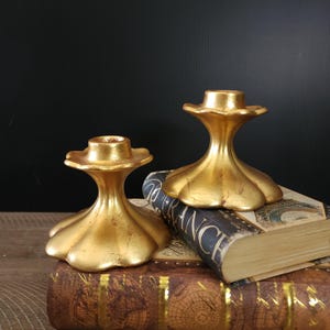 May include: Two gold-toned metal candle holders with a fluted design. The candle holders are sitting on top of a stack of books.