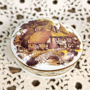 May include: A small, white, oval trinket box with a detailed landscape scene on the lid. The scene depicts a village with a bridge, houses, and trees in shades of brown, yellow, and green. The box has a gold trim.