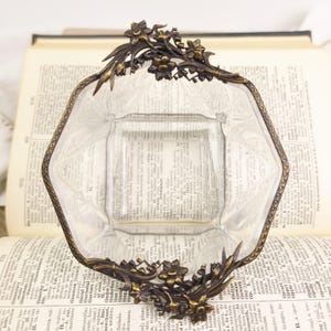 May include: Clear glass dish with an octagonal shape and a decorative bronze-colored metal frame. The frame features floral and leaf designs. The dish is sitting on top of an open book.