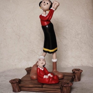 May include: A figurine of Olive Oyl and Popeye's baby, Swee'Pea, standing on a wooden platform. Olive Oyl is wearing a red shirt, black skirt, and yellow shoes. Swee'Pea is wearing a red onesie.