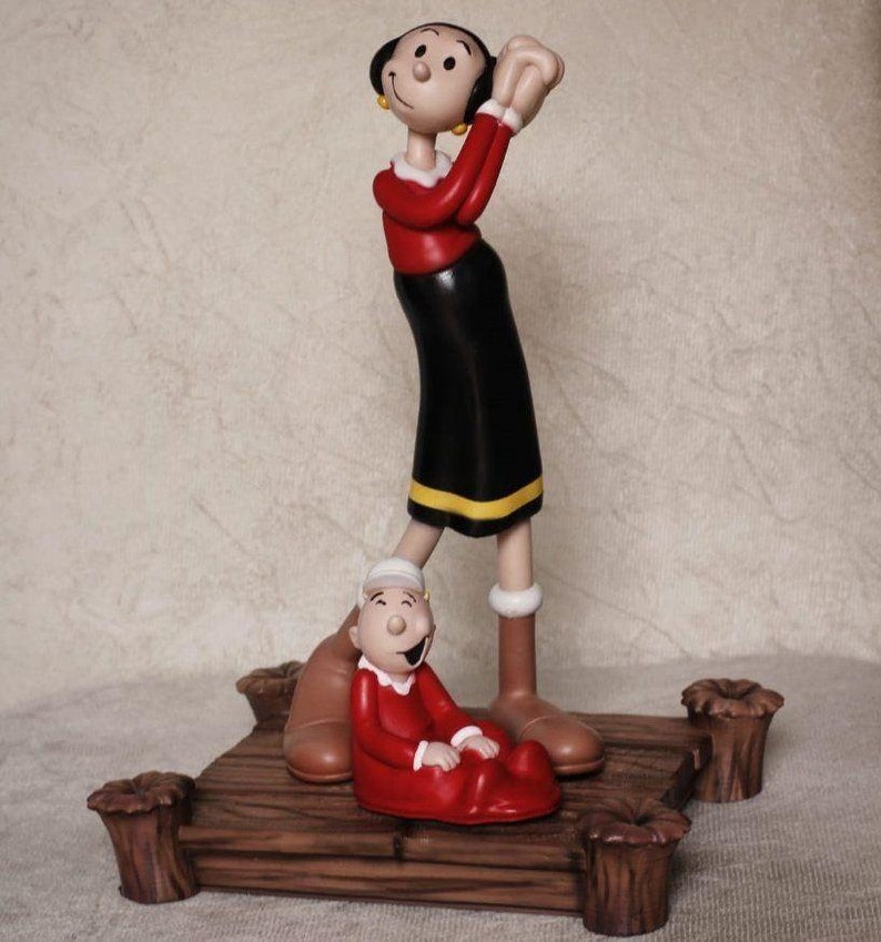 May include: A figurine of Olive Oyl and Popeye's baby, Swee'Pea, standing on a wooden platform. Olive Oyl is wearing a red shirt, black skirt, and yellow shoes. Swee'Pea is wearing a red onesie.