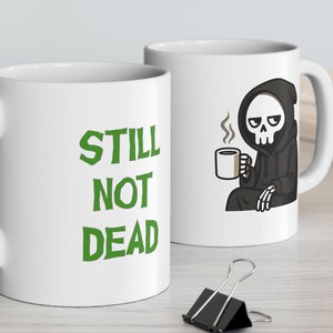 May include: Two white ceramic coffee mugs. One mug has the text "STILL NOT DEAD" in green. The other mug features a cartoon Grim Reaper character in a black robe, holding a coffee cup. The mugs are on a light-colored surface.