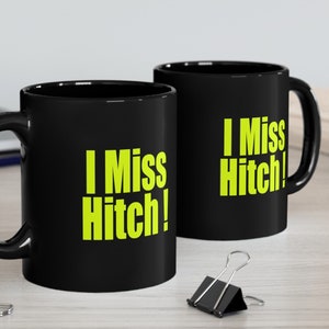 Puede incluir: Dos tazas de cerámica negra con una inscripción amarilla brillante que dice "I Miss Hitch!" Las tazas están sobre una superficie de madera con un lápiz y clips.