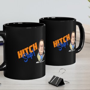 May include: Two black ceramic mugs with a cartoon image of a man and the text "Hitch Slap!" in orange and blue. The mugs are on a wooden table.