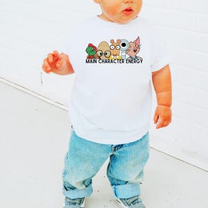 Könnte beinhalten: Ein weißes T-Shirt mit einem bunten Cartoon-Motiv und dem Text "MAIN CHARACTER ENERGY" in Schwarz. Das T-Shirt wird von einem Kind getragen, kombiniert mit blauen Jeans und grauen Turnschuhen.