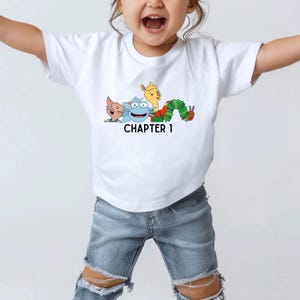 May include: A white t-shirt with a graphic of characters from the children's book The Very Hungry Caterpillar. The graphic includes a caterpillar, a llama, a cat, and a blue creature. The text "CHAPTER 1" is printed below the graphic.