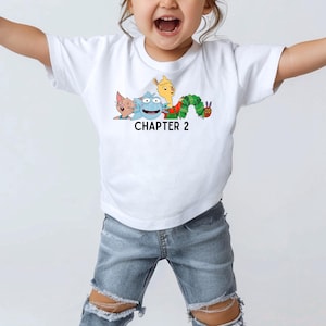 May include: White t-shirt featuring a graphic of characters from the book The Very Hungry Caterpillar. The graphic includes a blue monster, a yellow llama, a cat, and the caterpillar. The text "CHAPTER 2" is printed below the graphic.