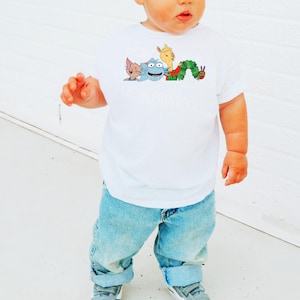 Op de afbeelding: Wit T-shirt met korte mouwen met een kleurrijke cartoonafbeelding van een kat, een blauw wezen, een lama en een rups. De tekst "MOROPONICO" staat onder de afbeelding. Het kind draagt lichtblauwe jeans en grijze sneakers.