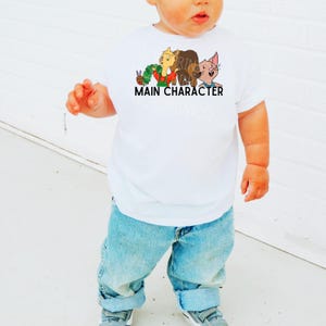 Op de afbeelding: Wit T-shirt met korte mouwen met een kleurrijke afbeelding van stripfiguren en de tekst "MAIN CHARACTER". Het shirt wordt gecombineerd met een blauwe spijkerbroek en grijze sneakers. Een casual outfit voor een kind.