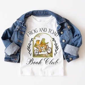 Könnte beinhalten: Weißes T-Shirt mit einer schwarzen und grünen Illustration von zwei Fröschen, die Fahrrad fahren. Die Illustration ist von einem Kranz aus grünen Blättern und dem Text "Frog and Toad Book Club" umgeben.