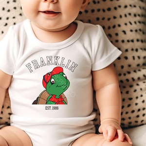 May include: White baby onesie featuring a cartoon turtle wearing a red cap and bow tie. The word "FRANKLIN" is arched above the turtle, with "EST. 1986" below. The onesie is short-sleeved and has a round neckline.