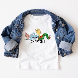 May include: White t-shirt with a graphic of characters from the book The Very Hungry Caterpillar. The graphic includes a blue monster, a yellow llama, and a green caterpillar. The text "CHAPTER 1" is printed below the graphic.