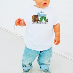 May include: A white t-shirt with a cartoon illustration of a caterpillar, a bear, and a blue character. The text "Main Character Energy" is printed below the illustration.