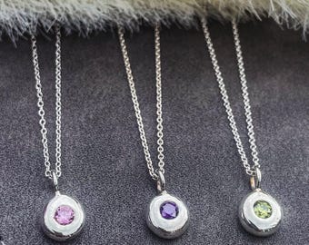 May include: Three silver necklaces with round pendants featuring a single gemstone. The pendants are silver with a smooth, rounded surface. The gemstones are pink, purple, and green.