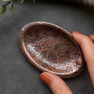 May include: An oval-shaped copper-toned dish featuring an embossed pattern of hummingbirds, tropical leaves, and various flowers. The surface has a metallic finish with dark recessed details that highlight the intricate botanical and avian motifs.