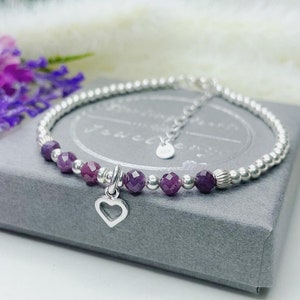 May include: A silver bracelet with purple beads and a small heart charm. The bracelet is on a grey background.