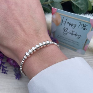 May include: A silver bracelet with small round beads and larger square beads. The bracelet is on a person's wrist. The card in the background says "Happy 70th Birthday".