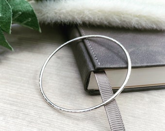 May include: A silver hammered bangle bracelet, lying on a gray book with a white ribbon bookmark.