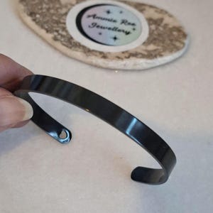 May include: A smooth, matte black metal cuff bracelet with a simple open-ended design. The bracelet features a small circular hole at one end of the band. The surface of the metal is solid black with a consistent finish throughout the entire piece.