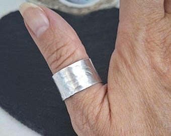 May include: A wide silver band ring featuring a hammered, textured finish. The metal surface has a reflective, metallic sheen with irregular indentations across the entire exterior, creating a light-catching, uneven appearance on the finger.