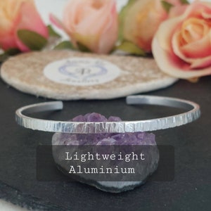 May include: A silver aluminium bangle bracelet with a hammered texture. The bracelet is lightweight and has a simple, modern design.