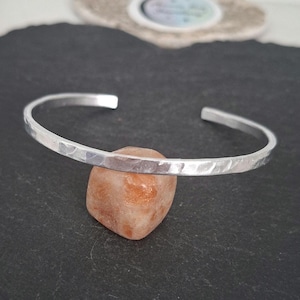 May include: A silver-coloured metal cuff bracelet with a hammered, textured surface. The bracelet is open-ended and rests on top of a small, smooth, orange-toned stone. The metal has a reflective finish that catches the light across its surface.