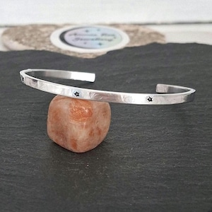 May include: A silver-coloured metal cuff bracelet with a hammered texture and small, engraved star motifs spaced along the outer band. The bracelet features an open design, allowing for adjustment, and has a polished, reflective finish.