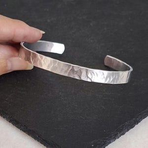 May include: A silver-coloured metal cuff bracelet featuring a hammered, textured surface. The bracelet has an open-ended design with a polished finish that reflects light across its uneven, indented pattern, creating a distinct visual appearance.