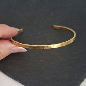 May include: A gold-coloured metal cuff bracelet with a hammered, textured surface. The bracelet features an open-ended design, allowing for adjustment. The metal has a reflective, polished finish that catches the light across its uneven surface.