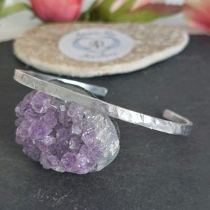 May include: A silver-toned metal cuff bracelet featuring a hammered texture on the band. One end of the open-style bracelet is adorned with a cluster of raw, deep purple amethyst crystals, showcasing natural jagged edges and a crystalline surface.