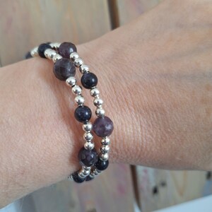 May include: A double-strand bracelet featuring alternating faceted purple gemstone beads and small, smooth, reflective silver-toned metallic beads. The design consists of two rows of beads wrapped around the wrist, showcasing a mix of dark and light purple tones.