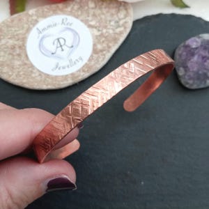 May include: A copper cuff bracelet featuring a repeating herringbone or chevron-style textured pattern across its outer surface. The metal has a warm, reddish-brown metallic finish with a polished sheen, and the bracelet has an open-ended design.