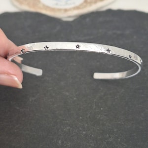 May include: A silver cuff bracelet featuring small star-shaped patterns. The bracelet has a textured finish and an open design. The stars are evenly spaced around the bracelet. The bracelet is made of a silver-colored metal.