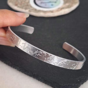 May include: A silver cuff bracelet featuring an engraved floral design. The open-ended bracelet is made of a shiny, metallic material. The floral pattern is etched into the surface, giving a delicate, textured look. The bracelet is displayed against a dark background.