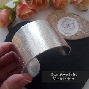 May include: A wide, silver cuff bracelet made from lightweight aluminium. The bracelet has a textured surface with a crisscross pattern. The text "Lightweight Aluminium" is visible in the image.