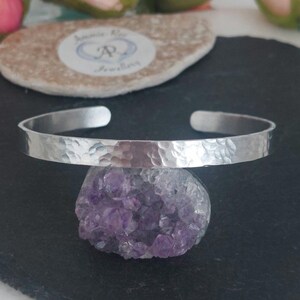 May include: A silver-coloured metal cuff bracelet with a hammered, textured finish. The bracelet is open-ended and rests on top of a cluster of purple amethyst crystals. The metal surface reflects light, highlighting the uneven, dimpled pattern.