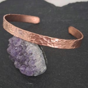 May include: A copper cuff bracelet featuring a hammered texture across its entire outer surface. The bracelet is an open-ended band design with a metallic reddish-brown finish, showcasing a consistent dimpled pattern throughout the metal band.