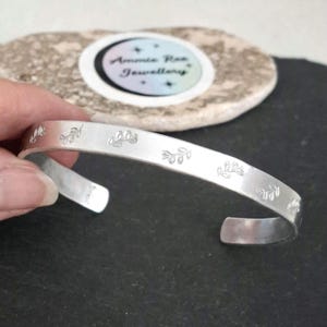 May include: A silver cuff bracelet featuring a leaf design. The open bracelet is held by a hand, with leaves spaced around the band. The bracelet is approximately 1.3 cm wide.