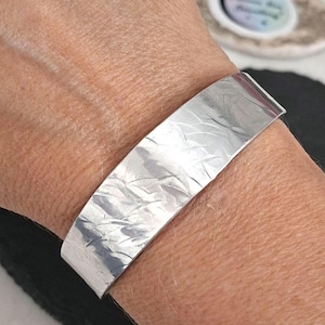 May include: A silver-toned metal cuff bracelet featuring a wide, flat band with a textured, hammered finish that creates a reflective, irregular pattern across the surface. The bracelet is worn around the wrist, showcasing its metallic sheen.