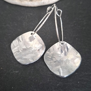 May include: A pair of silver earrings. Each earring has a textured, square pendant hanging from a long, curved wire. The pendants have a hammered look, reflecting light. The earrings are shown against a dark background.