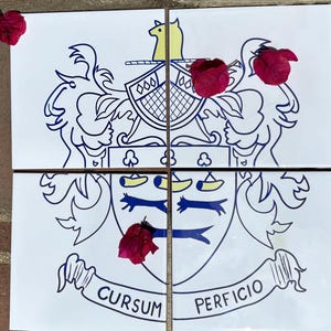 Könnte beinhalten: Eine weiße Keramikfliese mit einem blauen und gelben Wappen. Das Wappen zeigt einen Schild mit einem gelben Vogel, einem blauen und gelben Helmzier und einer blauen und gelben Schriftrolle mit dem Text "CURSUM PERFICIO".