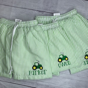 May include: Two pairs of green and white striped shorts with a green tractor embroidered on each pair. The shorts have a drawstring waist and the tractors have the names "Parker" and "Owen" embroidered on them.