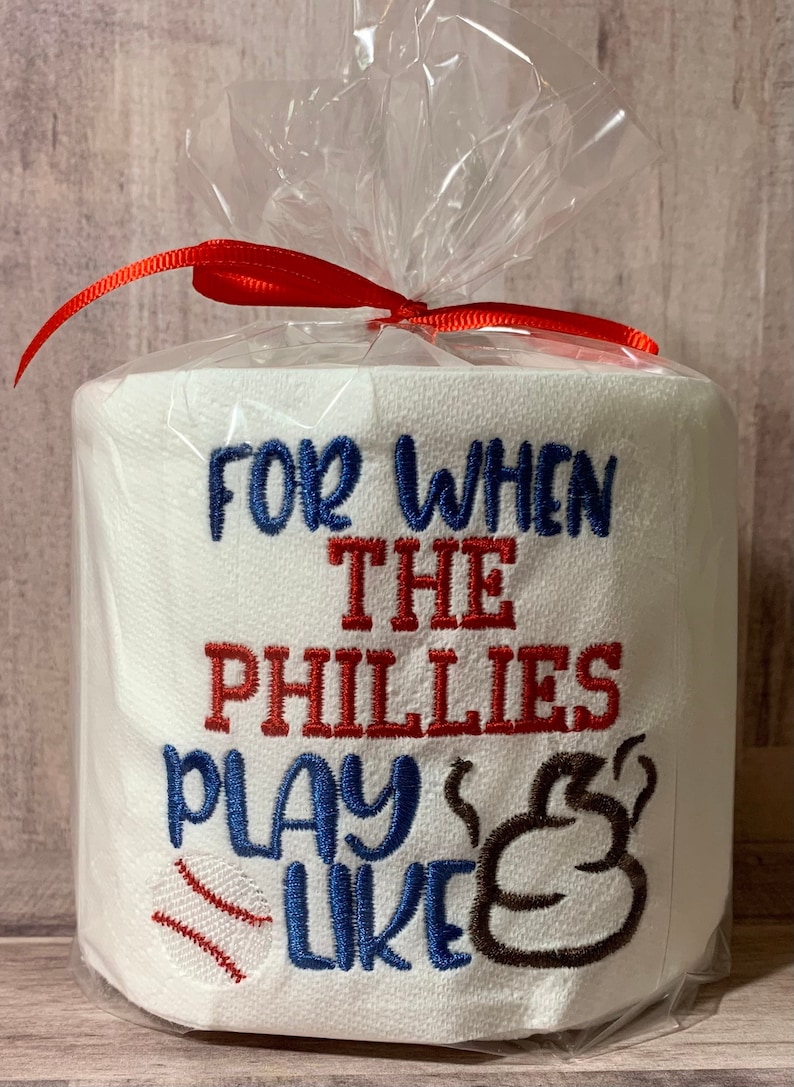 May include: A white toilet paper roll with the text "For when the Phillies play like" embroidered on it. The text is in blue and red, with a baseball and a brown poop emoji.